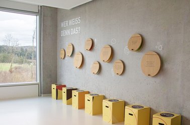 Moderne Wandgestaltung mit runden Holzplatten zum Aufklappen, darunter befinden sich gelbe Hocker.