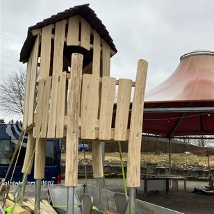 Neuer hölzerner Spielturm mit Dach auf einem Spielplatz, im Hintergrund ein großes rotes Sonnensegel und Tische