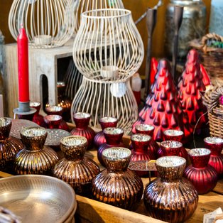 Verschiedene Weihnachtsdekorationen auf einem Marktstand darunter rote Kerzen, goldene Schalen, rote und goldene Kugeln sowie weiße Laternen aus Metall