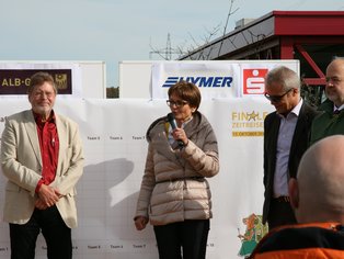 Vier Personen stehen vor einem Stapel weißer Kartons, eine Frau spricht in ein Mikrofon.