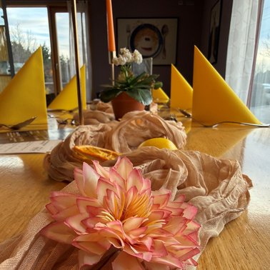 Tischdekoration mit rosa Blume, beigem Stoff, Zitrone und getrockneter Orangenscheibe, im Hintergrund gelb gefaltete Servietten und Kerzenhalter mit weißen Blumen