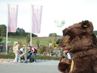 Menschen gehen auf einem Weg im Freien, im Vordergrund eine Person im Bärenkostüm