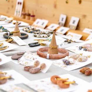 Schmuckstücke wie Ringe, Ohrringe und Armbänder auf einem hellen Verkaufstisch mit Holzständern im Hintergrund