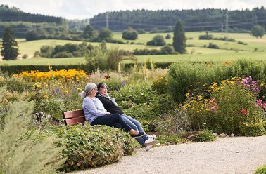 Zwei Frauen sitzen auf einer Bank in einem blühenden Garten mit grünen Hügeln im Hintergrund
