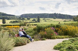 Zwei Frauen sitzen auf einer Bank in einem blühenden Garten mit grünen Hügeln im Hintergrund
