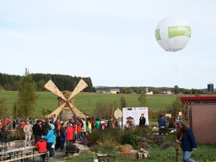 Menschenmenge bei einem Fest mit einem großen hölzernen Windrad und einem weißen Heißluftballon am Himmel in ländlicher Umgebung
