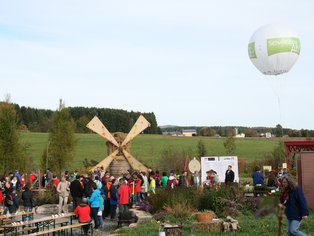 Menschenmengen bei einer Veranstaltung mit einer Luftballonfahrt und einer Windmühle im Hintergrund.