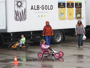 Kinder fahren mit Karts auf einem Parkplatz vor einem ALB-GOLD Anhänger.