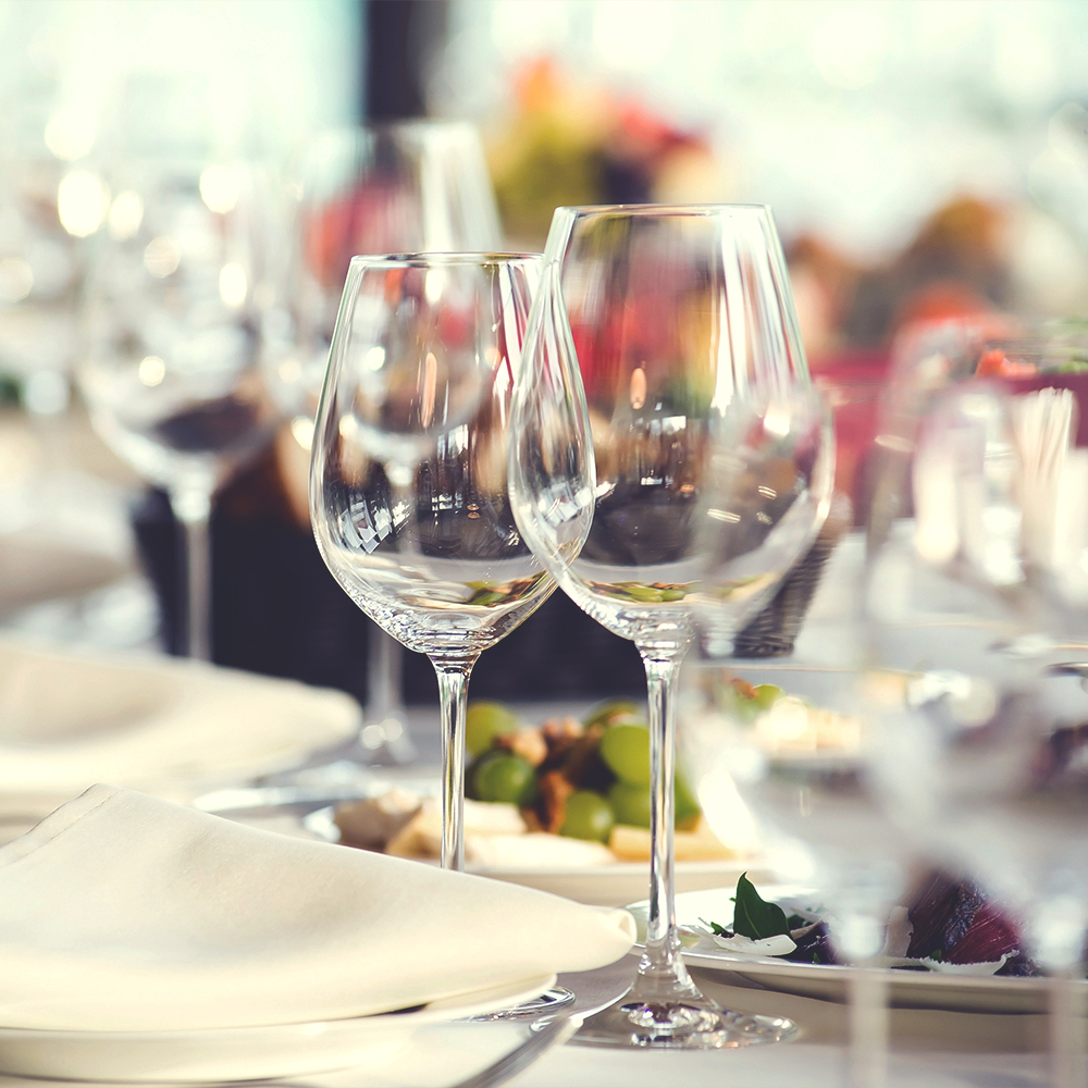 Weingläser auf gedecktem Tisch mit weißen Tellern und unscharfem Hintergrund mit Blumen und weiteren Gläsern