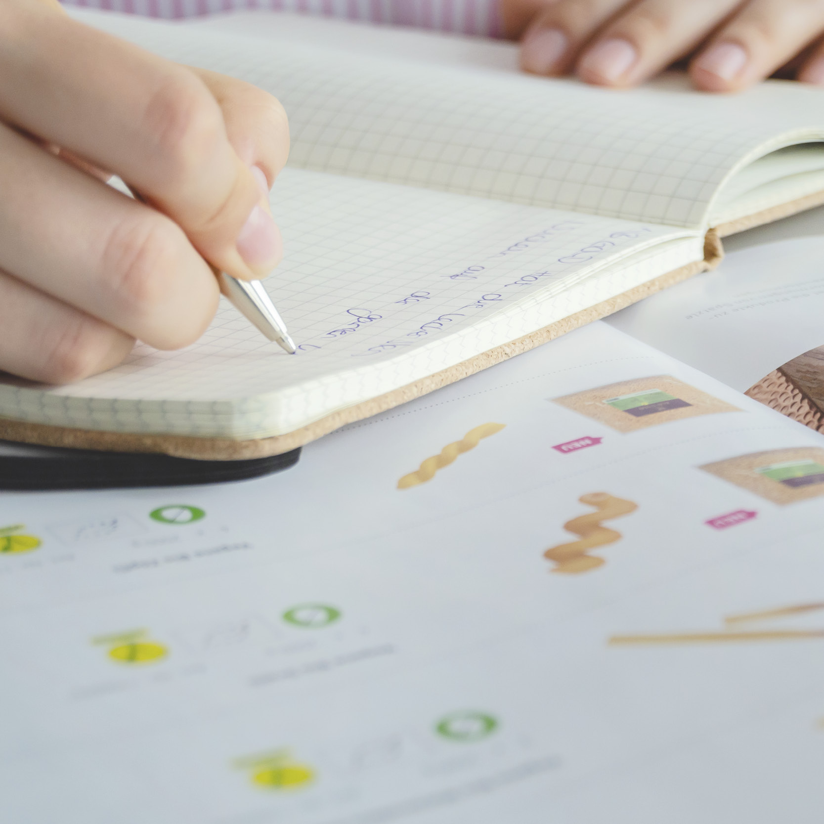Hand schreibt mit silbernem Kugelschreiber in kariertes Notizbuch, daneben liegen geöffnete Bücher mit Abbildungen und Texten.