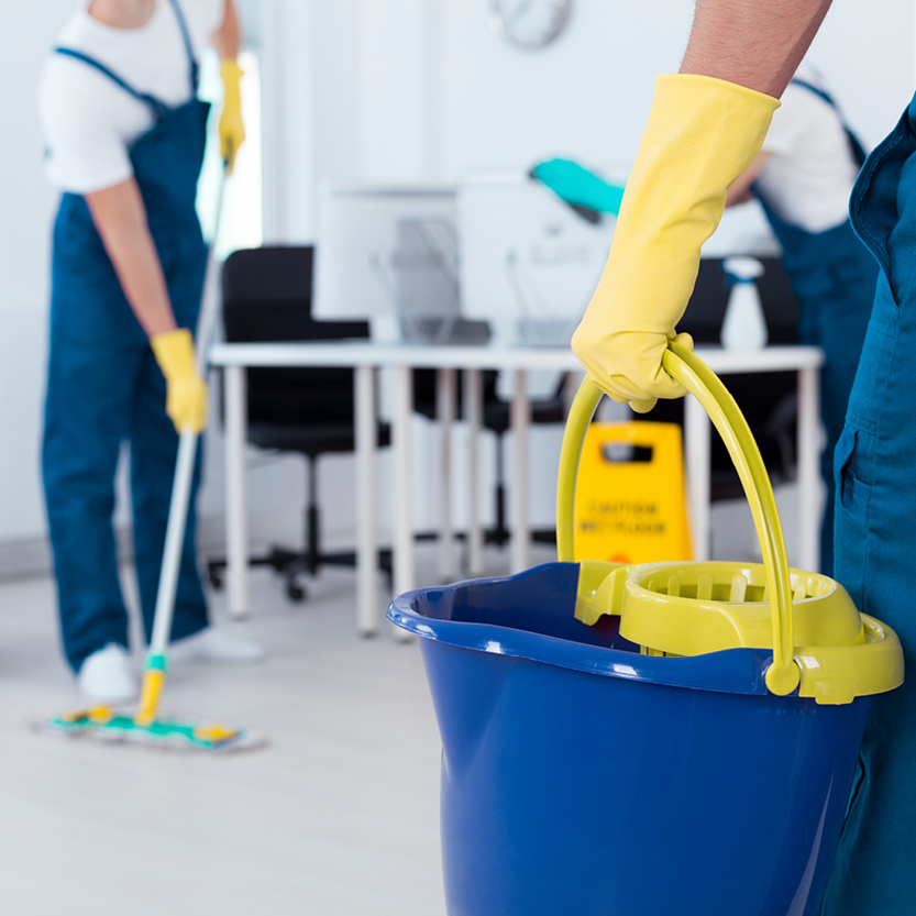 Zwei Reinigungskräfte in Arbeitskleidung und gelben Handschuhen reinigen einen Büroraum, eine Person hält einen blauen Wischeimer